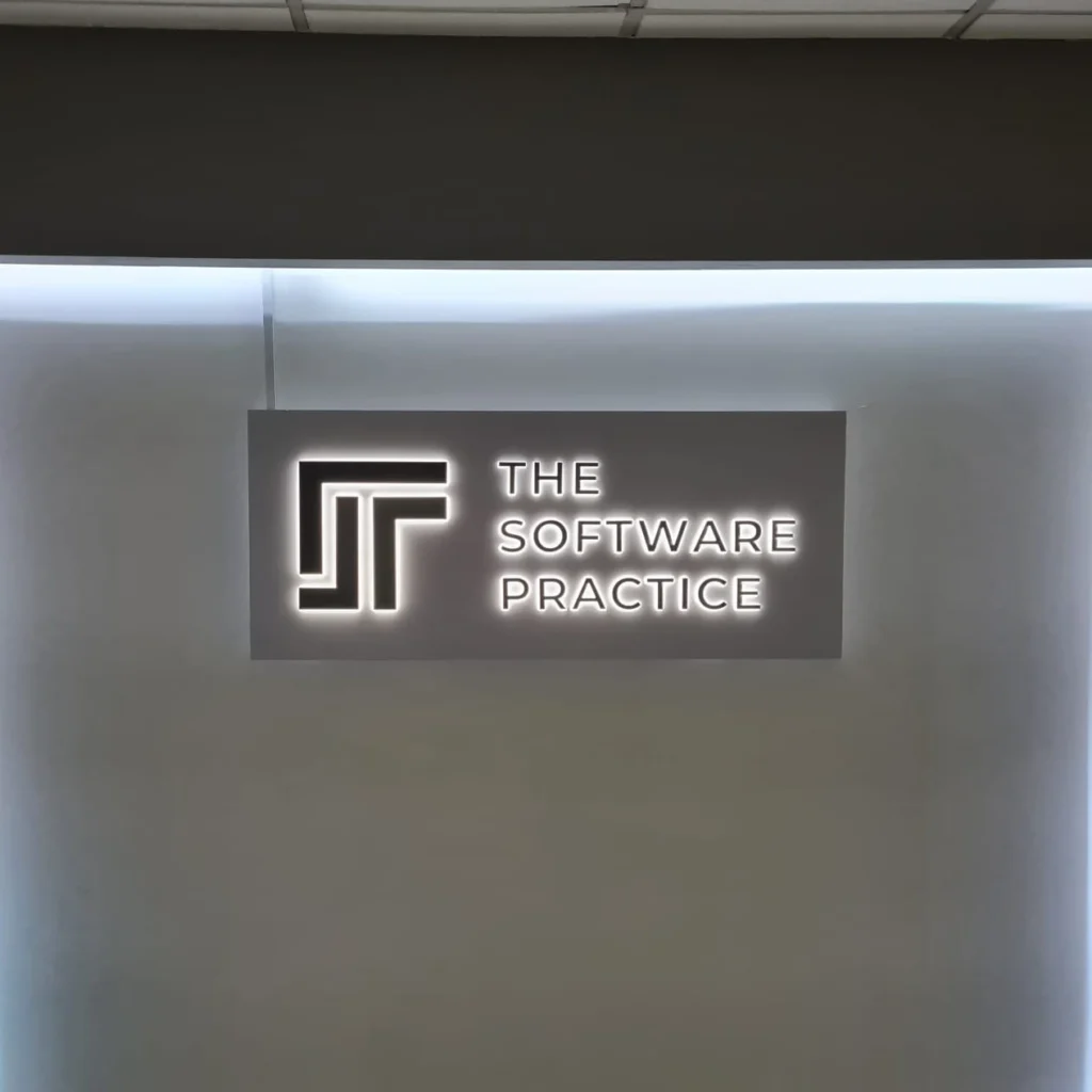 A backlit 3D boxup office reception sign displaying a geometric logo and the text "THE SOFTWARE PRACTICE" inside a white illuminated wall recess.