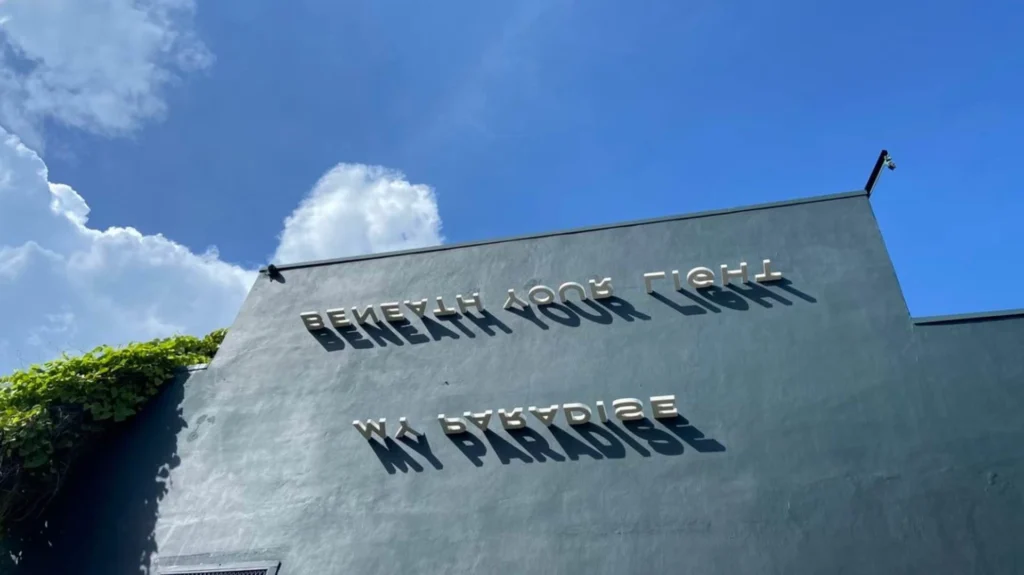 Outdoor 3D metal signage reading "BENEATH YOUR LIGHT MY PARADISE" mounted on a dark grey concrete wall under a bright blue sky