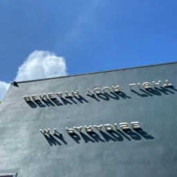Outdoor 3D metal signage reading "BENEATH YOUR LIGHT MY PARADISE" mounted on a dark grey concrete wall under a bright blue sky