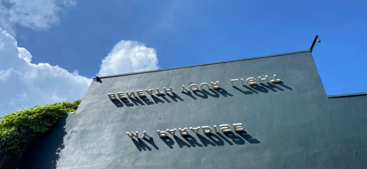 Outdoor 3D metal signage reading "BENEATH YOUR LIGHT MY PARADISE" mounted on a dark grey concrete wall under a bright blue sky