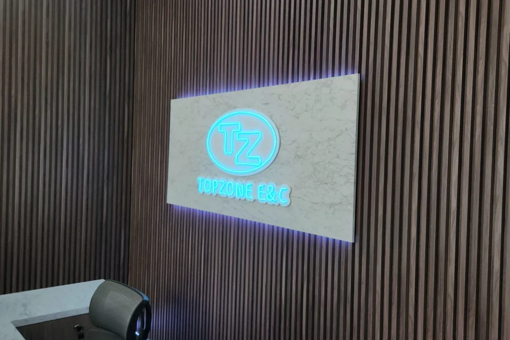 A glowing cyan neon corporate logo sign for "TOPZONE E&C" mounted on a marble plaque against a vertical wood slat office wall