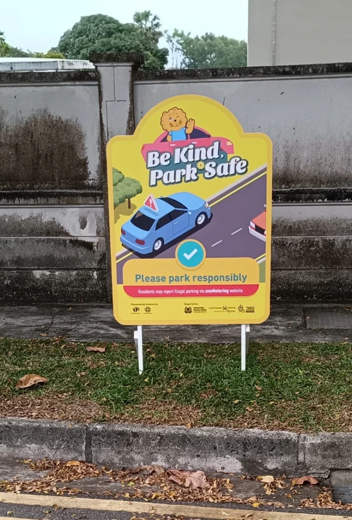 Bright yellow roadside signboard with bold graphics and text reading Be Kind Park Safe in Singapore.