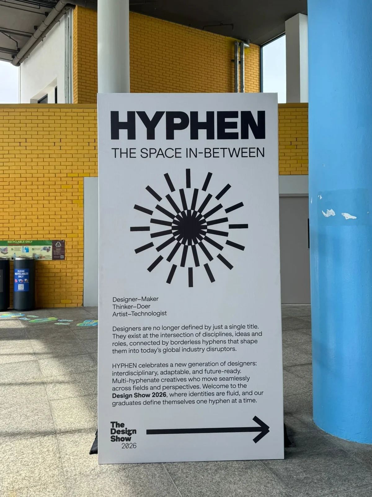 A tall, vertical white floor sign for "The Design Show 2026" titled "HYPHEN THE SPACE IN-BETWEEN"